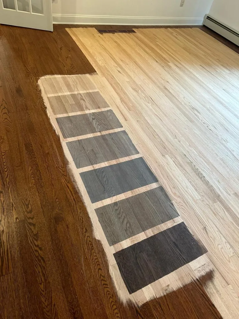 View across the room showing the taped grid of stain samples over a section that transitions from a light raw wood (ready for stain) to a darker finished wood. This highlights the comparative nature of hardwood floor color sampling.